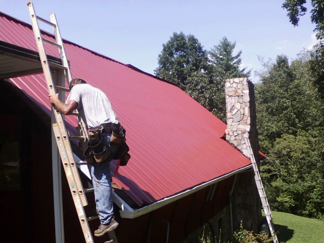Roofer on a ladder beside a red metal roof and stone chimney — Soffit & Fascia Repair by Roofing Brothers in Oxon Hill